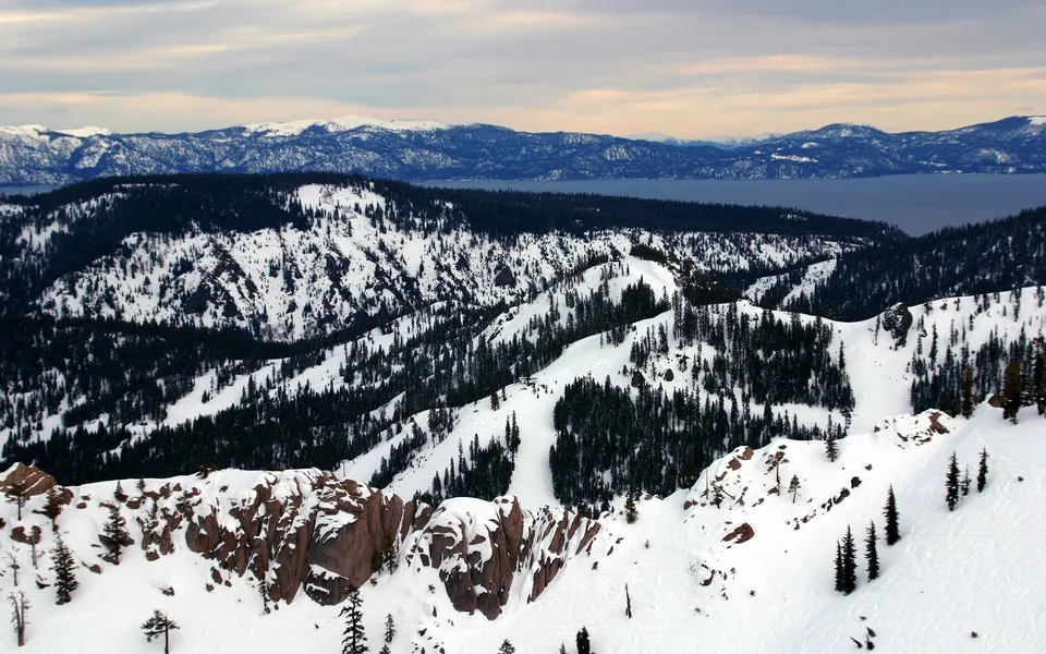 A wide view of snow-covered mountains and pine trees with a large lake in the background under a cloudy sky.