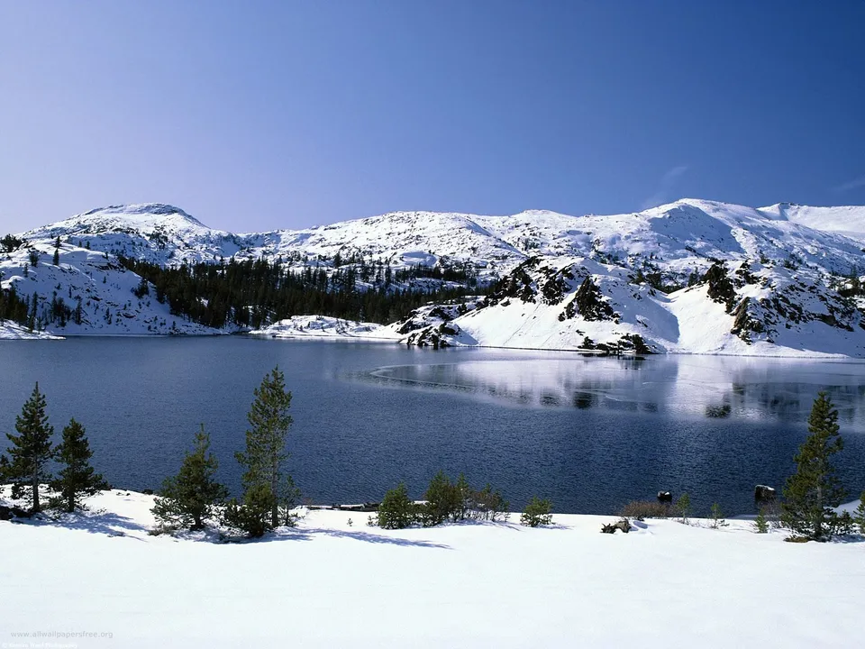 A wide, calm lake reflects snow-covered mountains and evergreen trees under a clear blue sky.