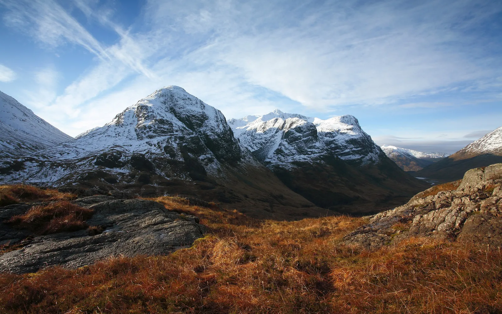 Snow-capped mountains rise above a valley with dry grass in the foreground under a blue sky with wispy clouds.