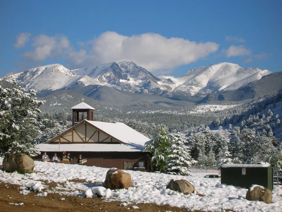 A snow-covered lodge sits in front of majestic snow-capped mountains under a blue sky with clouds.