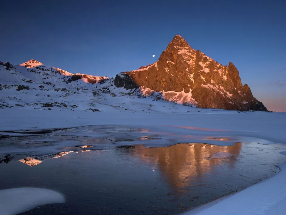 A snow-covered mountain peak is illuminated by the golden light of sunrise, with a reflection in a partially frozen body of water.