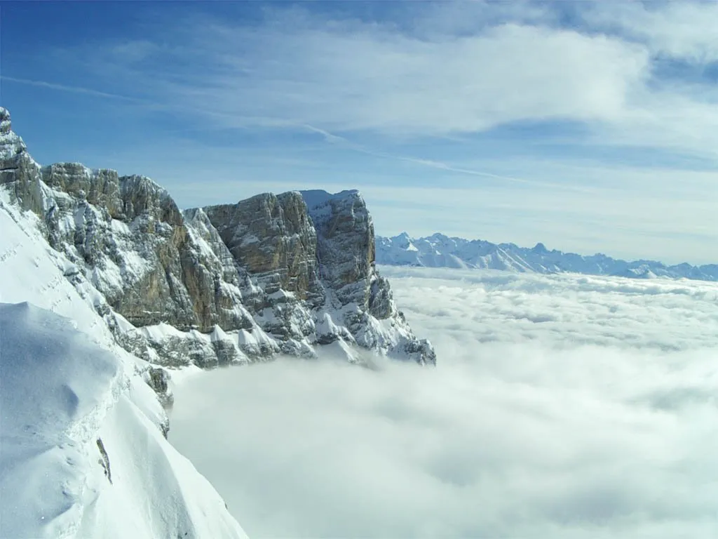 A vast expanse of snow-covered mountains rises above a thick blanket of clouds under a clear blue sky.