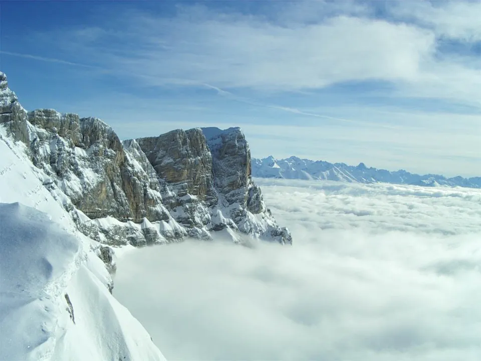 A vast expanse of snow-covered mountains rises above a thick blanket of clouds under a clear blue sky.