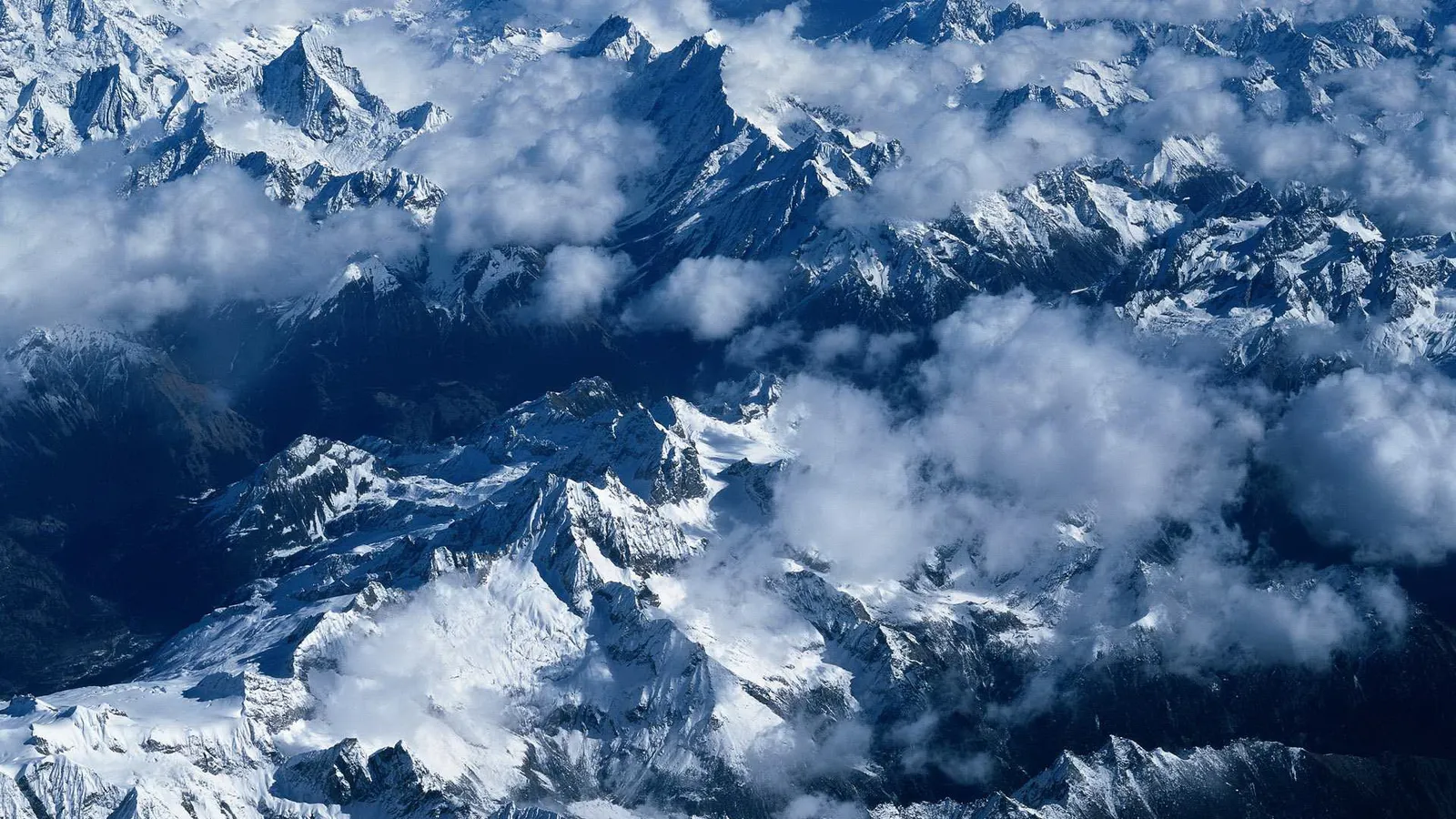 An aerial view of a vast mountain range covered in snow, with clouds drifting between the peaks.