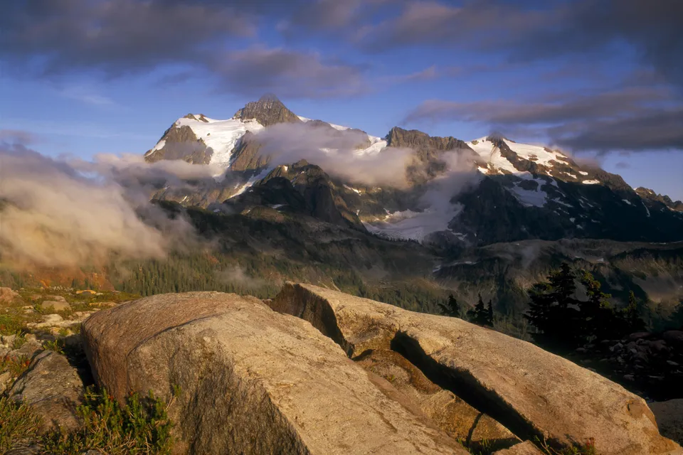 Snow-capped mountain peaks emerge from swirling clouds, with large rocks in the foreground.
