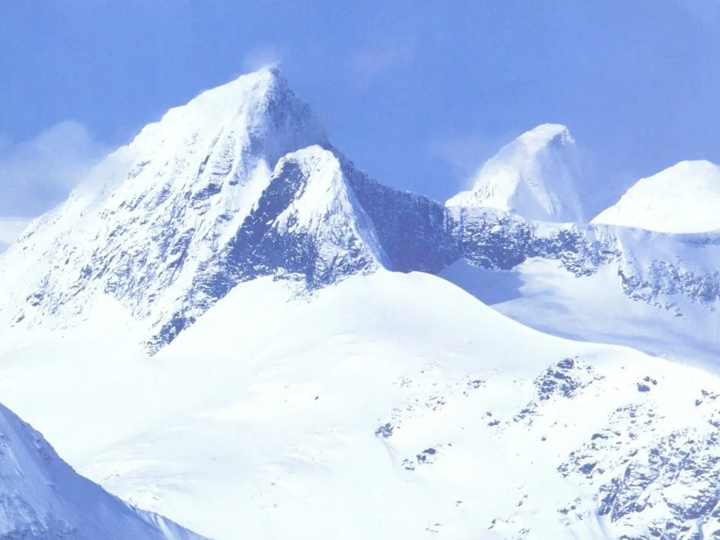 A wide shot of snow-covered mountain peaks under a bright blue sky with some wispy clouds.
