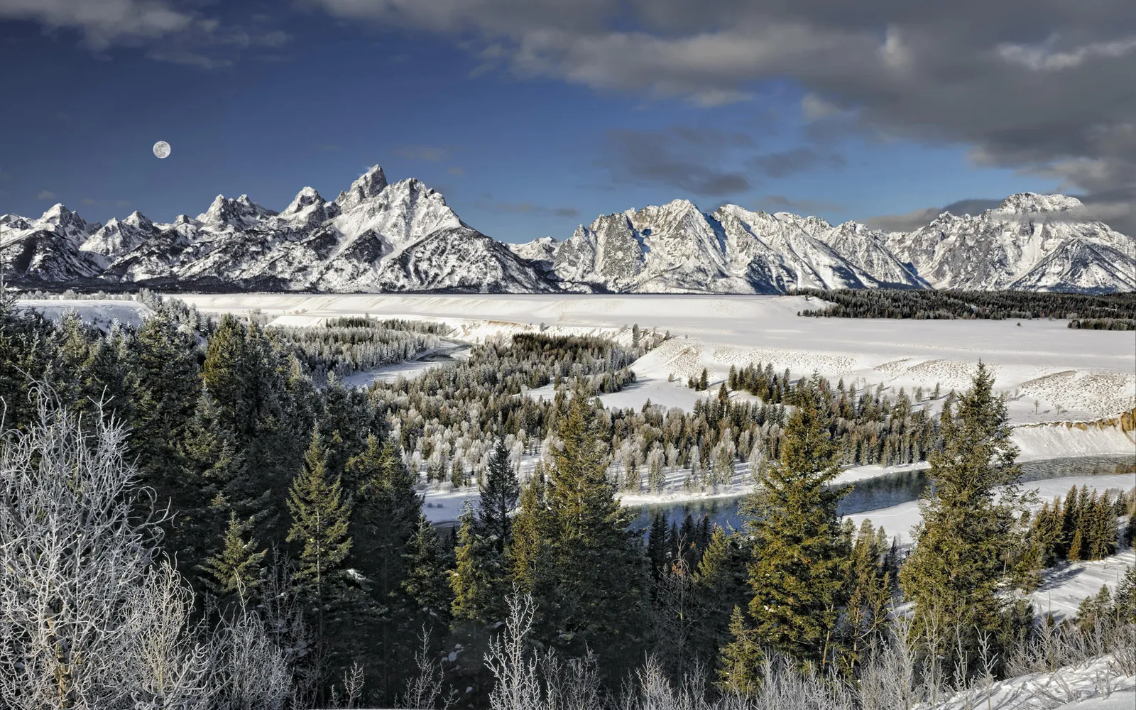 A vast, snow-covered mountain range is seen under a blue sky with a visible moon and foreground trees.