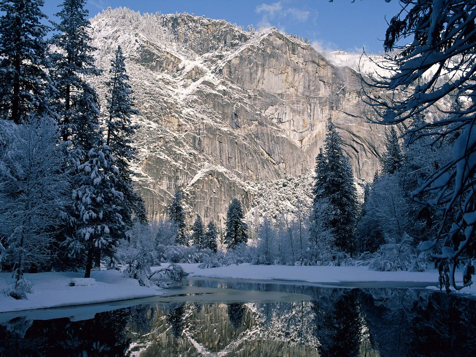 A snow-covered mountain looms over a partially frozen river, with pine trees lining the banks.