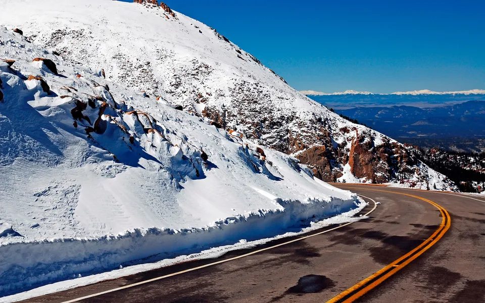 A winding asphalt road with yellow lines curves through a snow-covered mountain landscape under a clear blue sky.