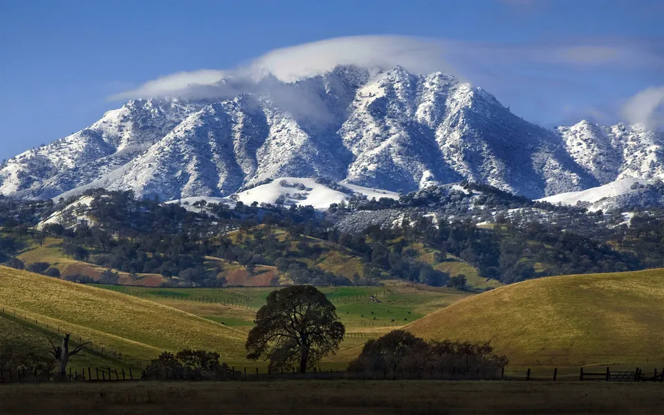 A majestic snow-covered mountain range looms over verdant, rolling hills under a bright blue sky with wispy clouds.
