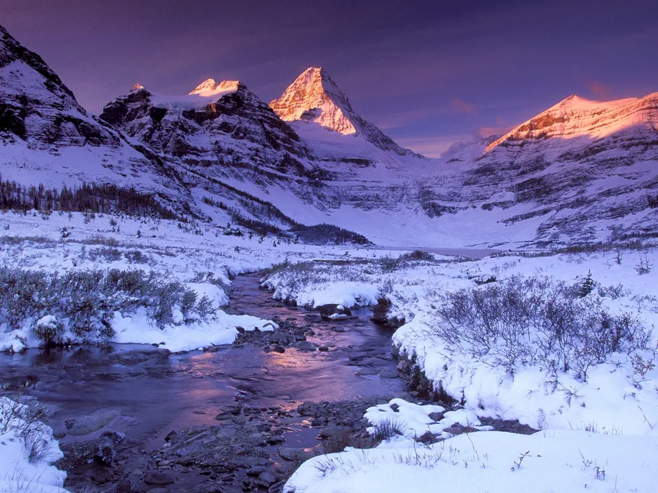 A snow-covered mountain valley with a stream flowing through it, illuminated by sunrise.
