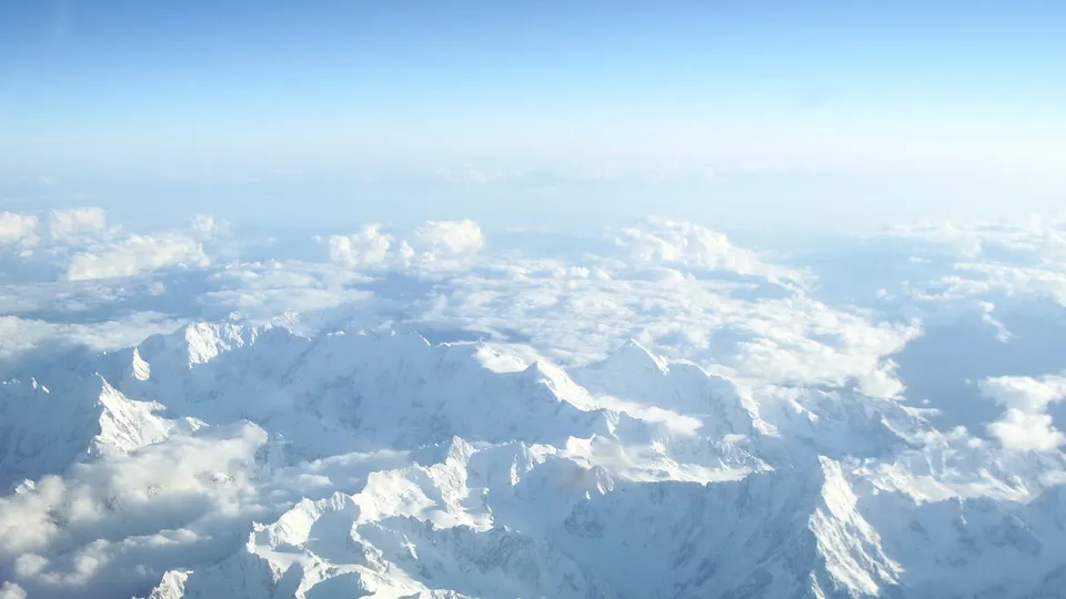 Aerial view of vast snow-covered mountain ranges peeking through fluffy white clouds under a clear blue sky.