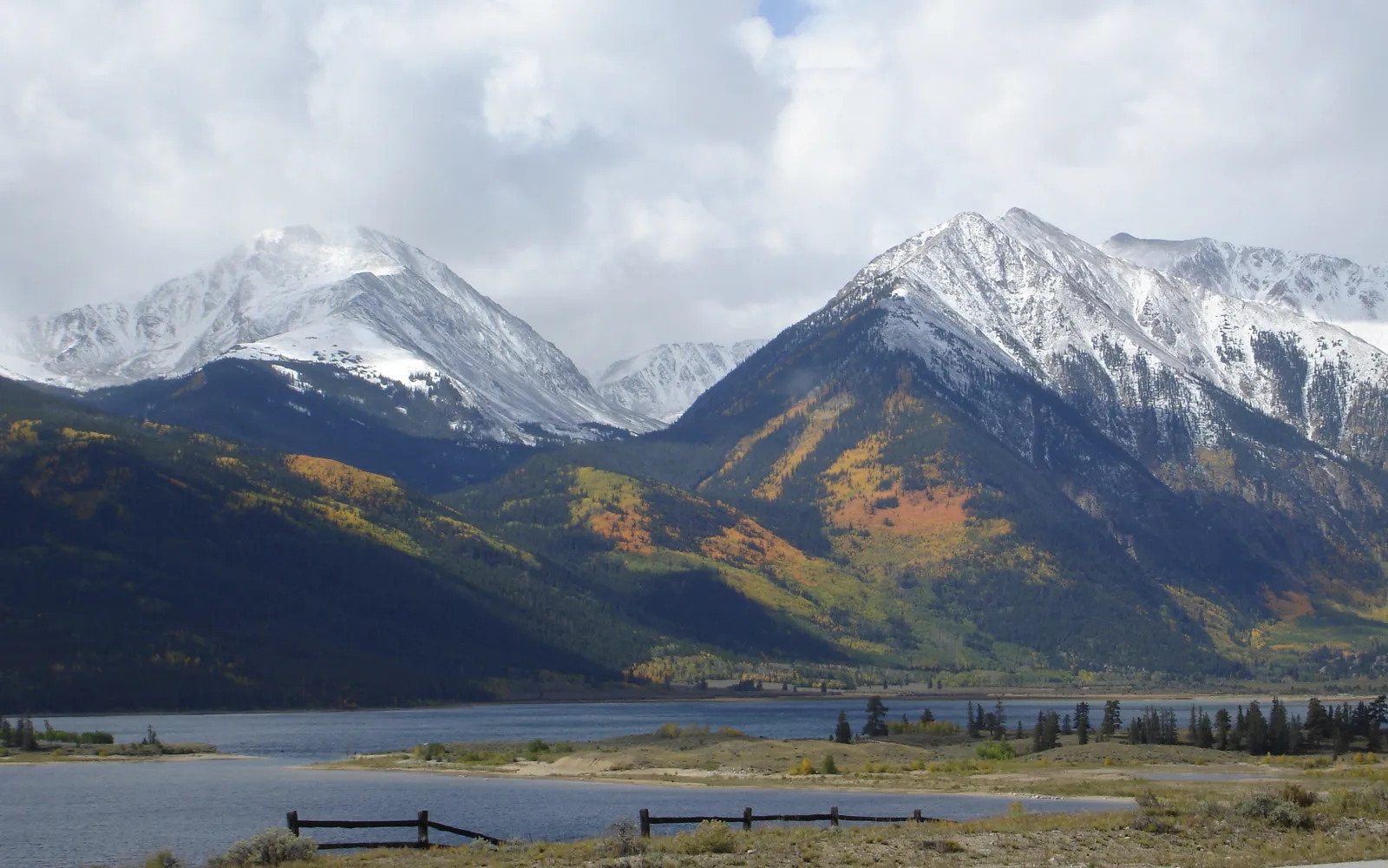 Snow-capped mountains rise above a lake, with hillsides showing autumn colors and dark green trees.