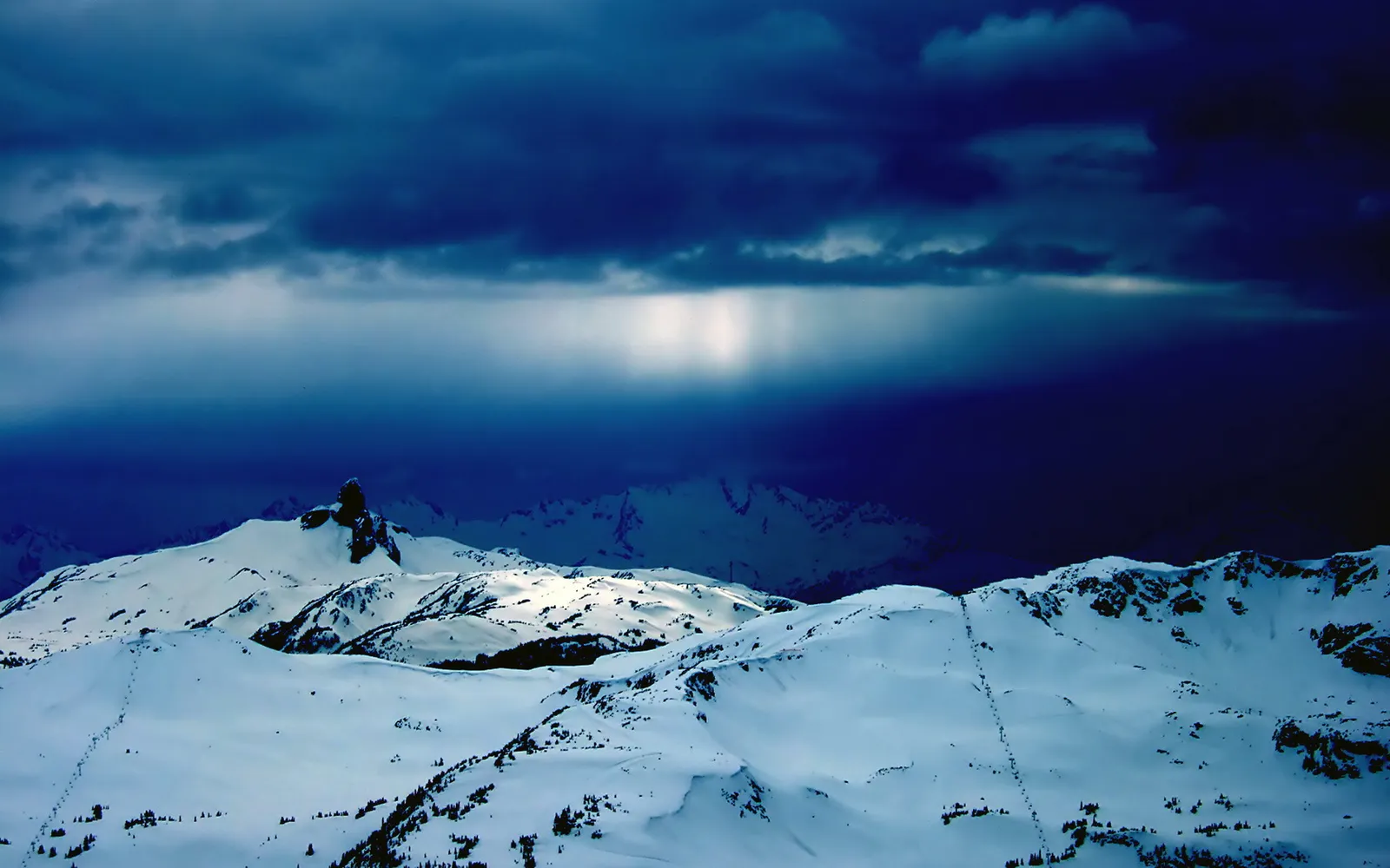 Snow-covered mountains under a dark, stormy sky with a beam of light breaking through.