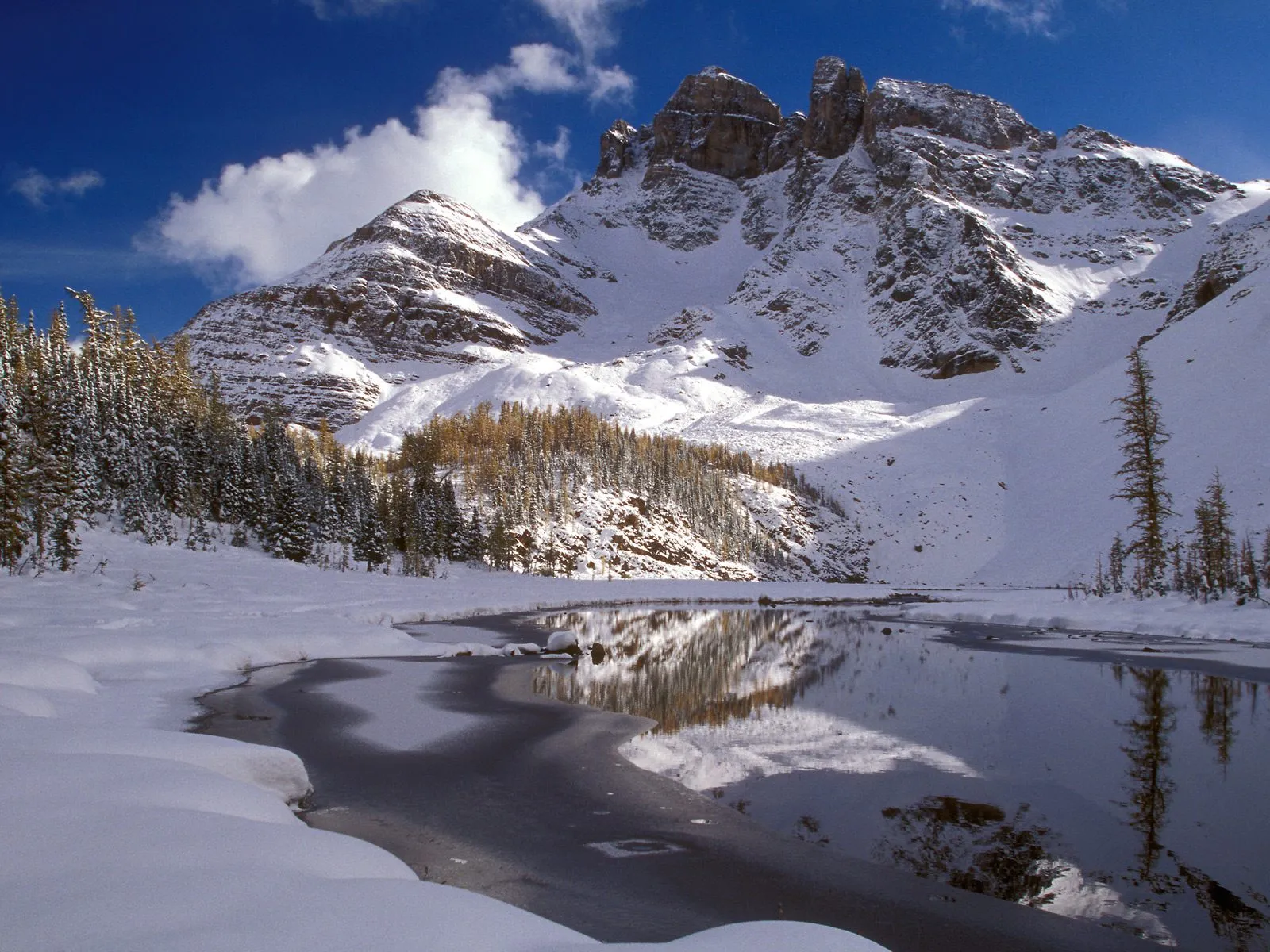 A snow-covered mountain range is reflected in a partially frozen lake under a bright blue sky with clouds.