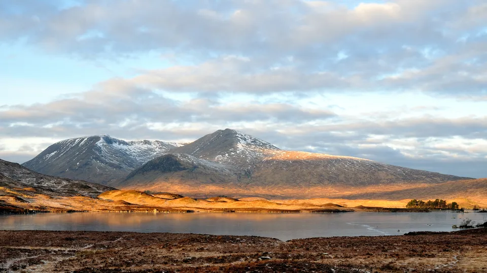 Snow-capped mountains are illuminated by warm golden light, with a calm lake in the foreground under a cloudy sky.