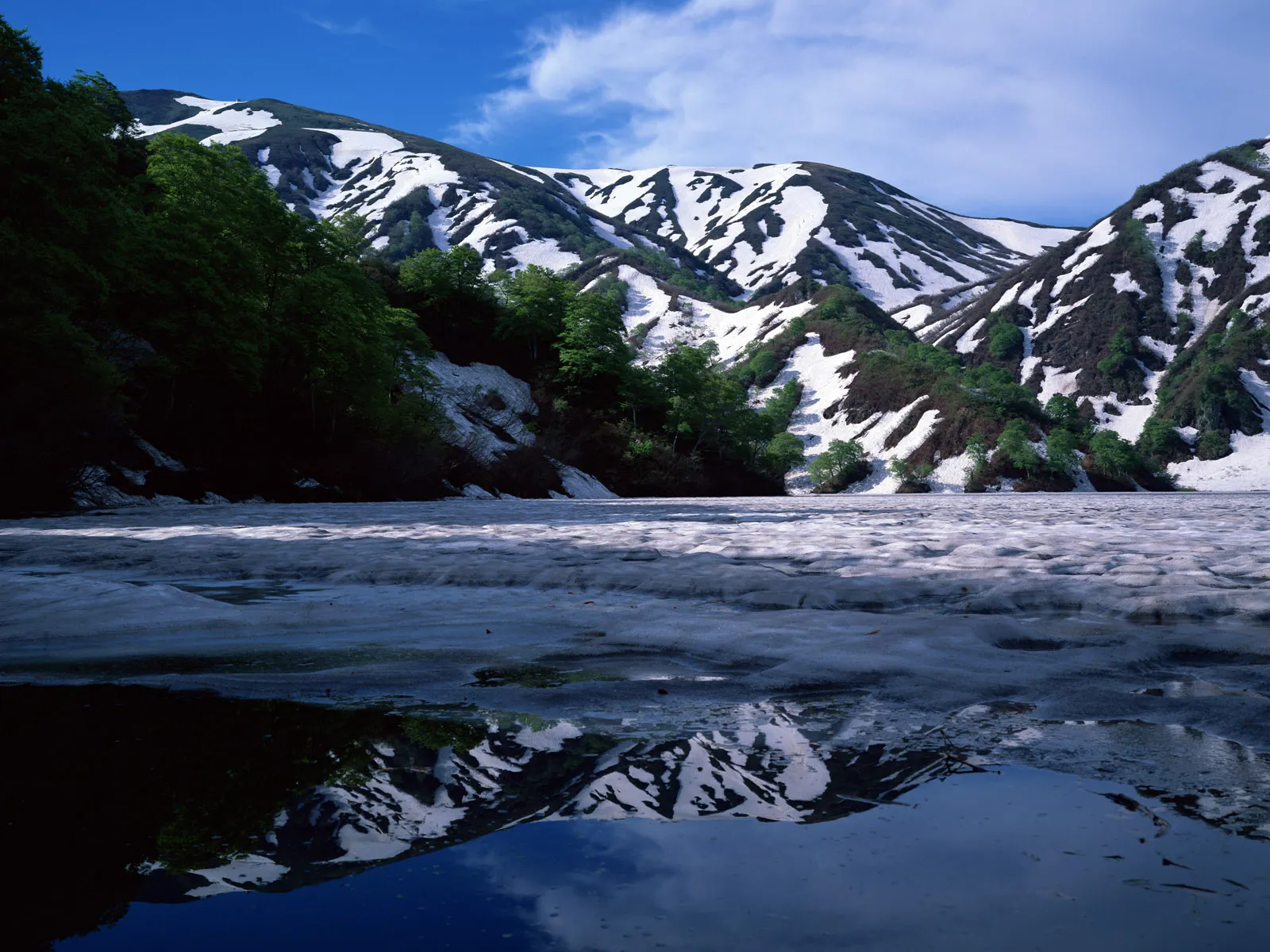 Snow-covered mountains with green trees reflected in a partially frozen body of water under a blue sky.