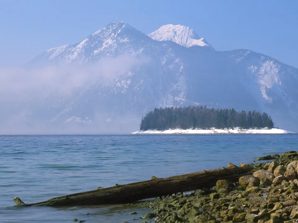 A calm lake with a snow-covered island and mountains in the background.