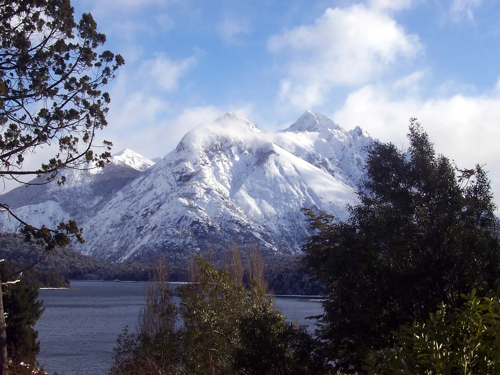 Snow-covered mountains rise majestically above a dark lake, with trees in the foreground and clouds in the sky.