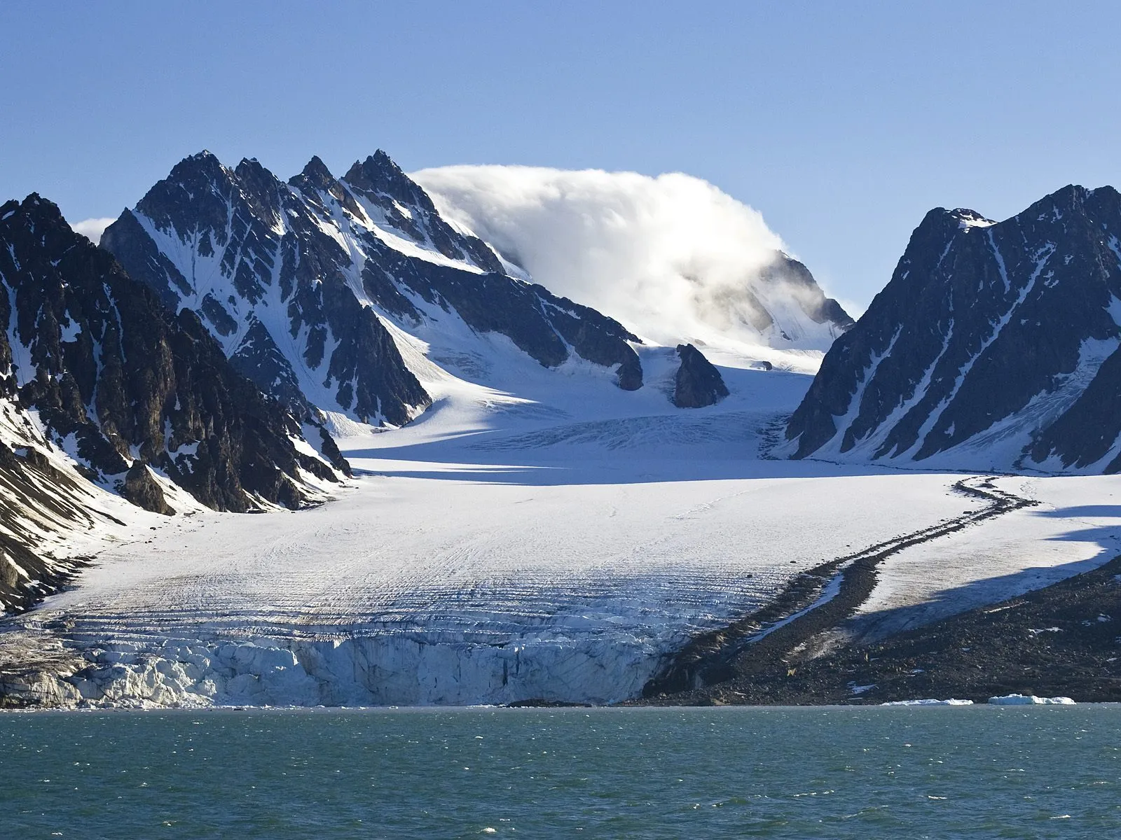 A vast glacial landscape with snow-covered mountains meeting a dark, choppy ocean under a clear blue sky.