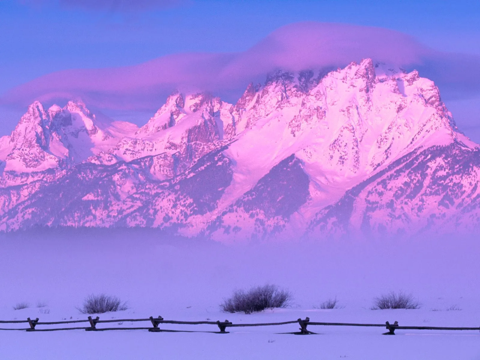 Snow-covered mountains glow with pink and purple light under a hazy sky, with a wooden fence in the foreground.
