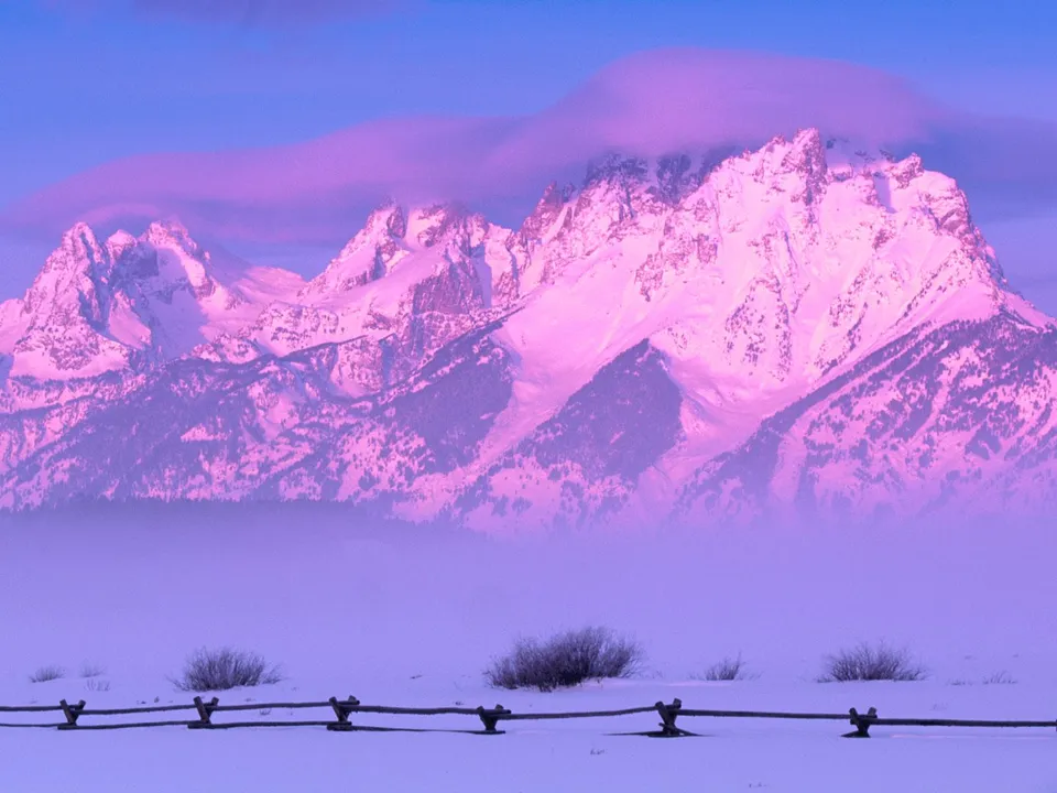 Snow-covered mountains glow with pink and purple light under a hazy sky, with a wooden fence in the foreground.