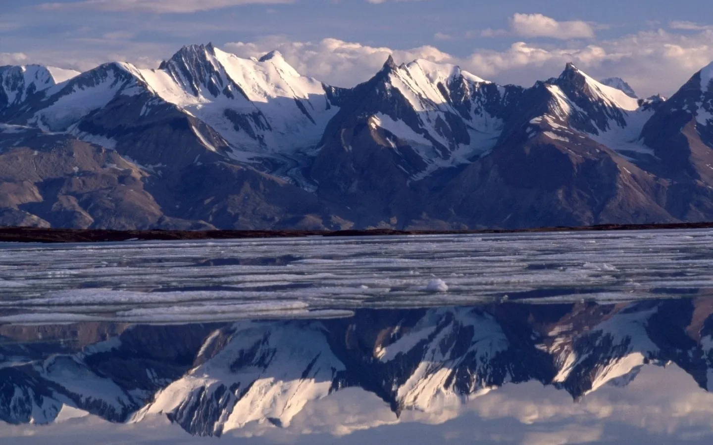 Snow-capped mountain range reflected in a partially frozen body of water under a cloudy sky.