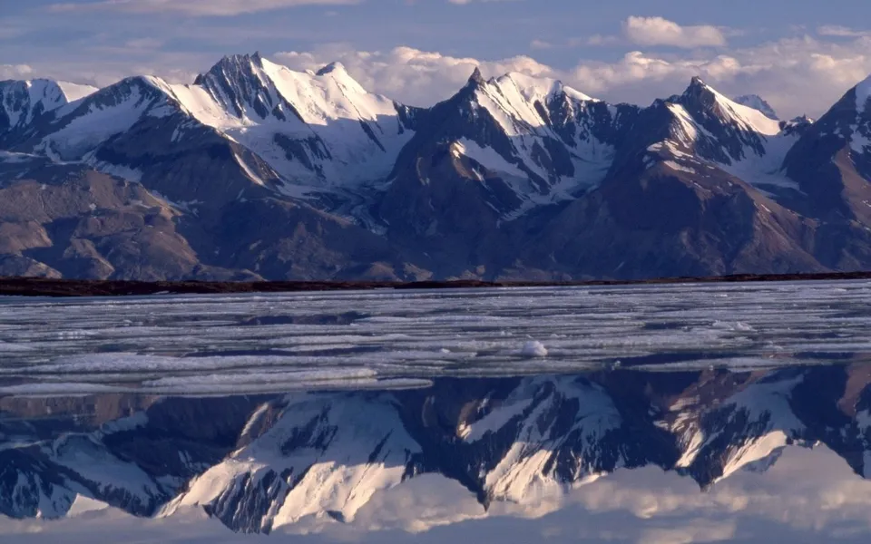 Snow-capped mountain range reflected in a partially frozen body of water under a cloudy sky.