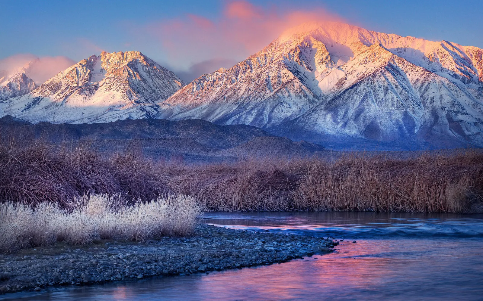 Snow-capped mountains bathed in pink sunrise light, with a calm river in the foreground and dry reeds.