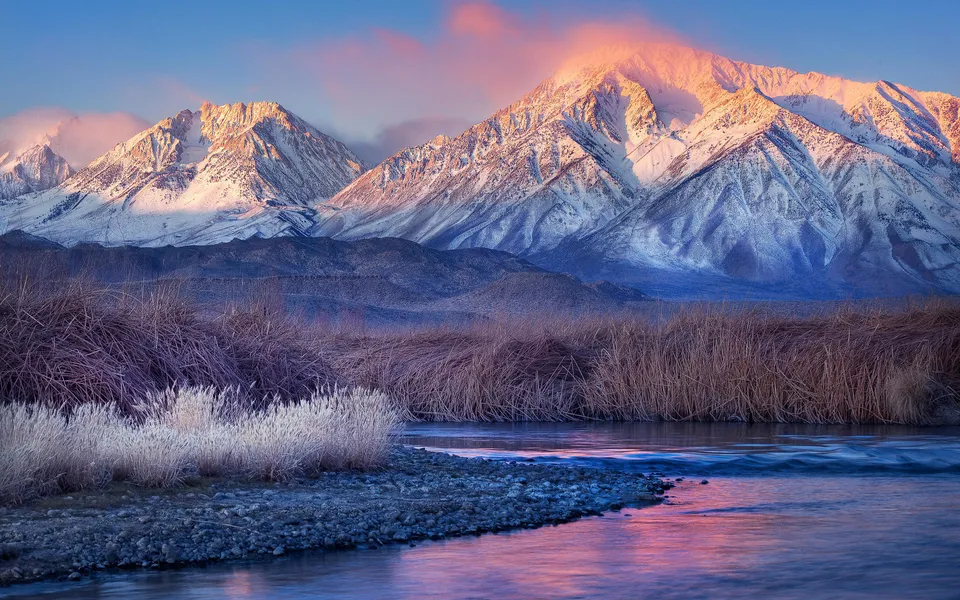 Snow-capped mountains bathed in pink sunrise light, with a calm river in the foreground and dry reeds.