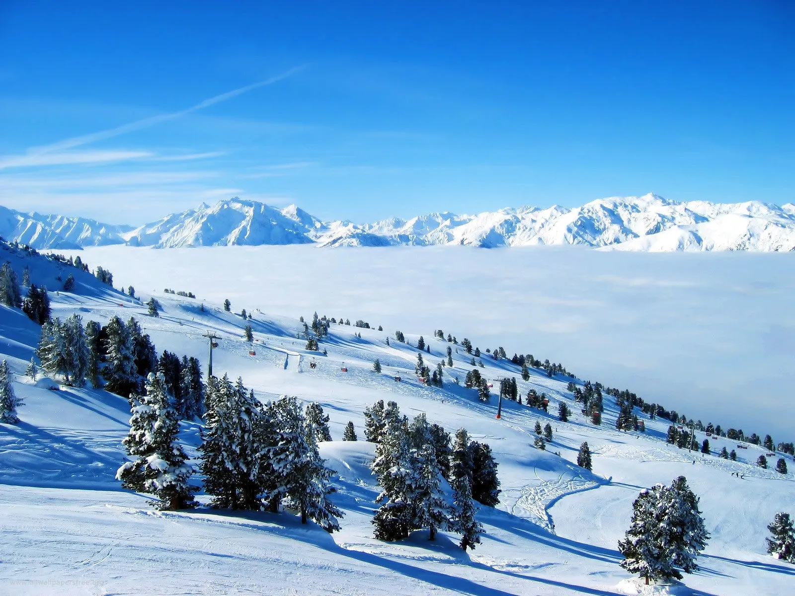 A vast snowy mountain range is visible above a thick layer of clouds, with a ski slope in the foreground.