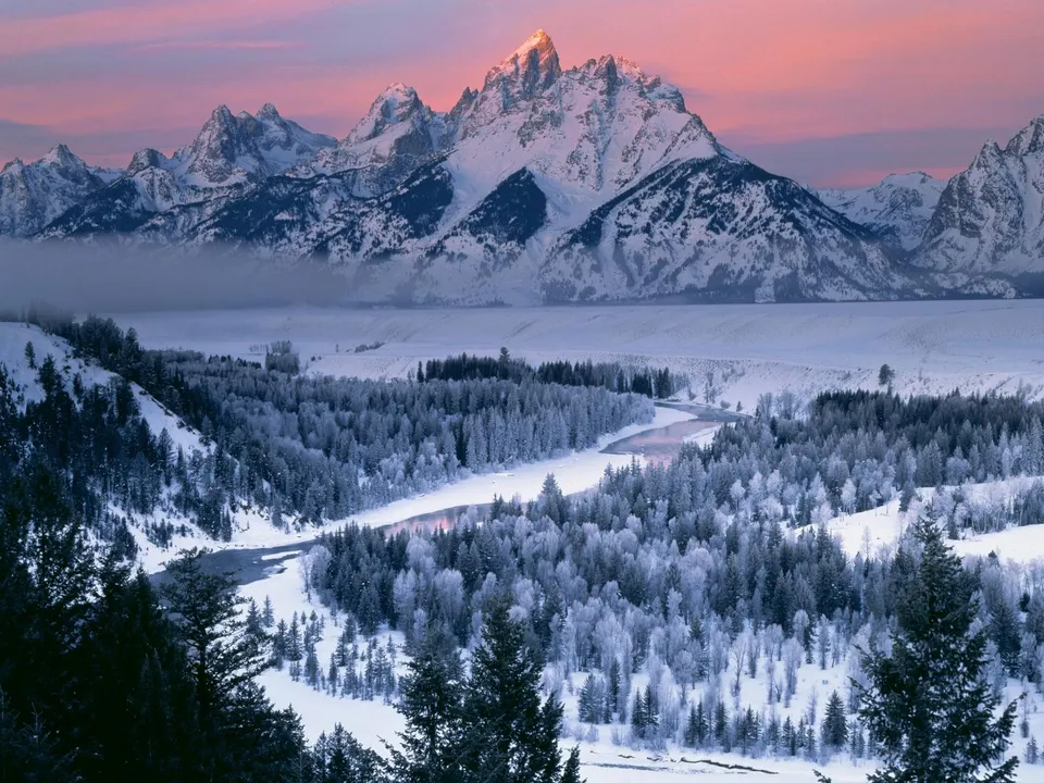 A vast snowy mountain range is illuminated by the soft pink light of sunrise, with a frosted forest and river in the foreground.