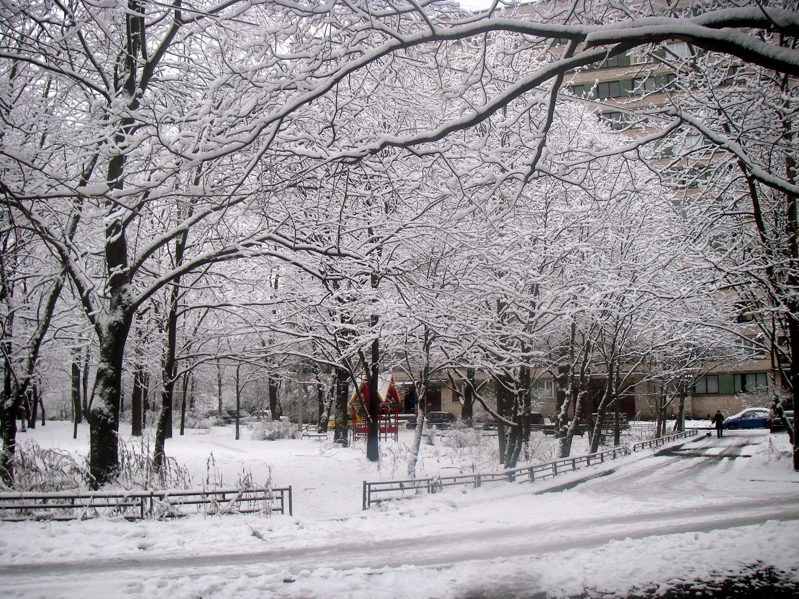 A park covered in snow with trees and a building in the background.