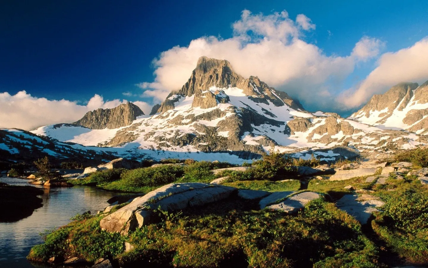 A snow-dusted mountain peak rises above lush green vegetation and a calm body of water under a blue sky.