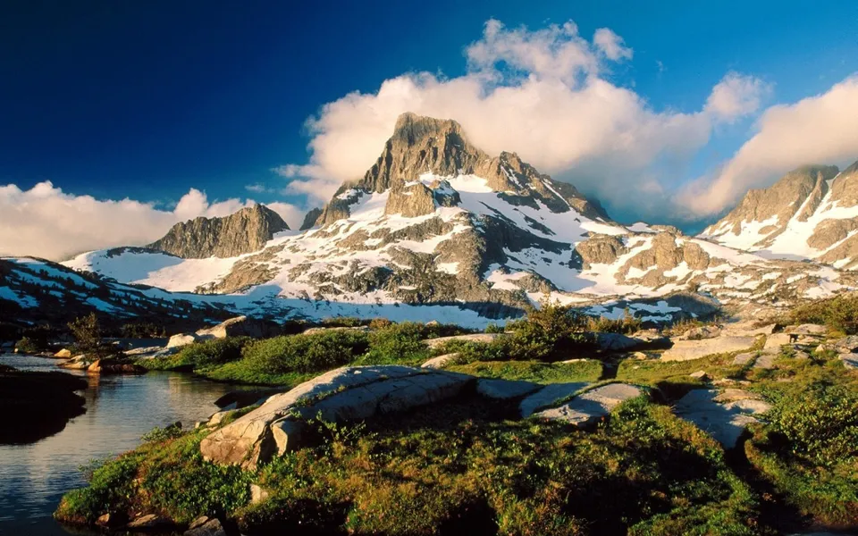 A snow-dusted mountain peak rises above lush green vegetation and a calm body of water under a blue sky.