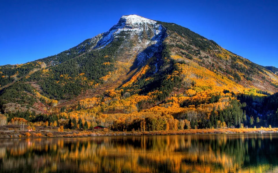 A snowy mountain peak overlooks a lake reflecting brilliant yellow and orange autumn colors.