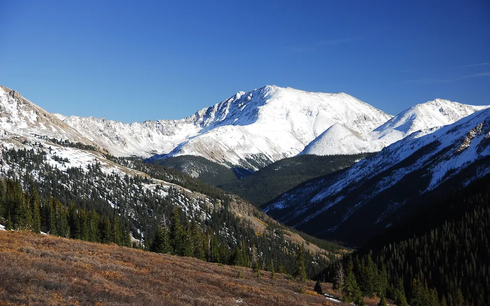 Snowy mountain peaks rise above a valley filled with dark evergreen forests and golden autumn foliage.