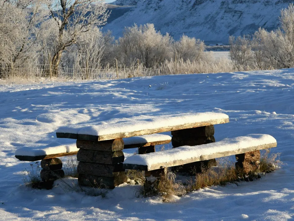 A wooden picnic table covered in snow sits in a snowy field with frosted trees and mountains in the background.