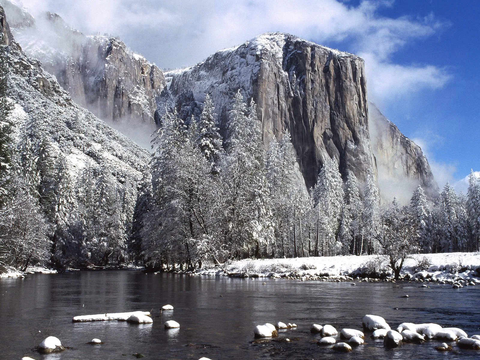 A wide river flows in the foreground, with snow-covered trees and a large, sheer cliff face in the background under a blue sky.