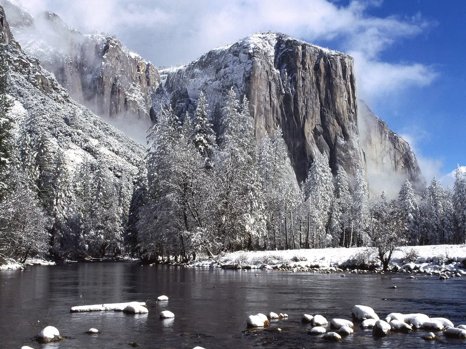A wide river flows in the foreground, with snow-covered trees and a large, sheer cliff face in the background under a blue sky.