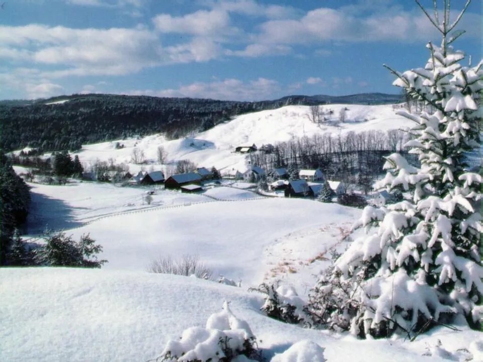 A small village is nestled in a valley surrounded by snow-covered hills under a bright blue sky.