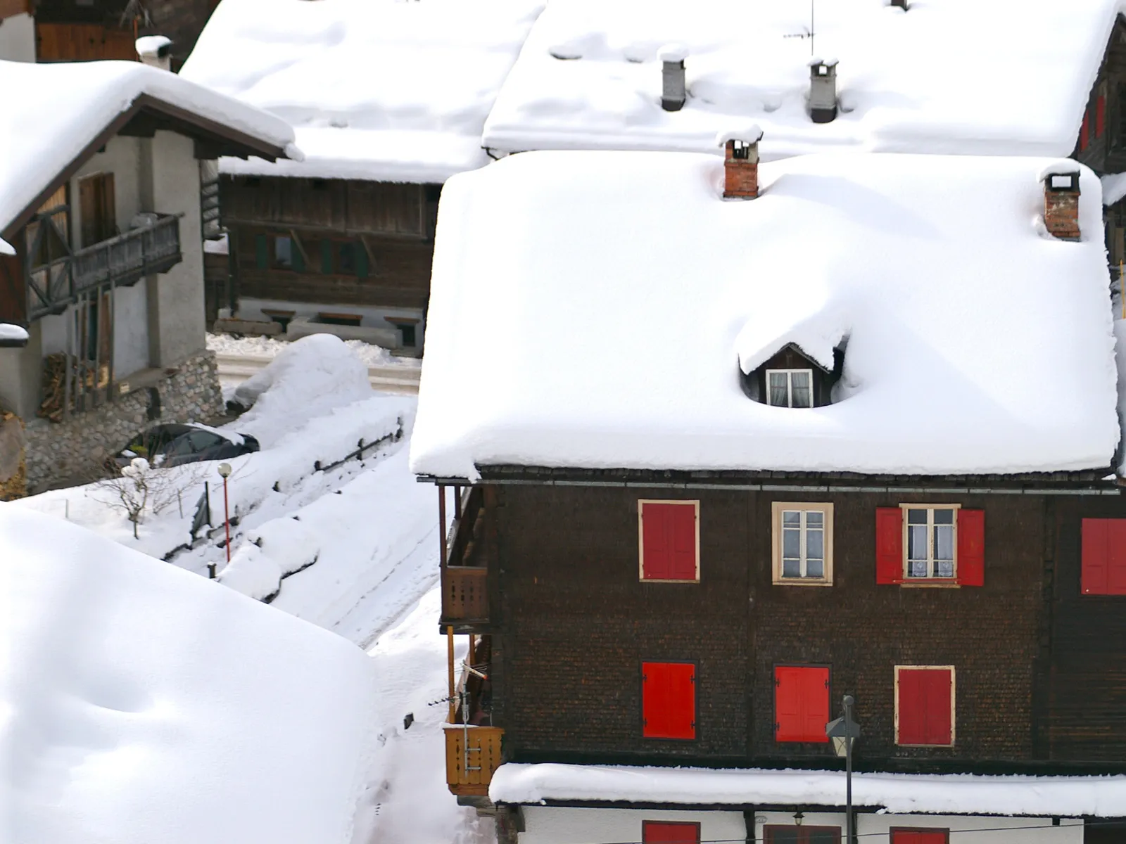 Traditional houses with red shutters are covered in thick snow, with chimneys poking through.