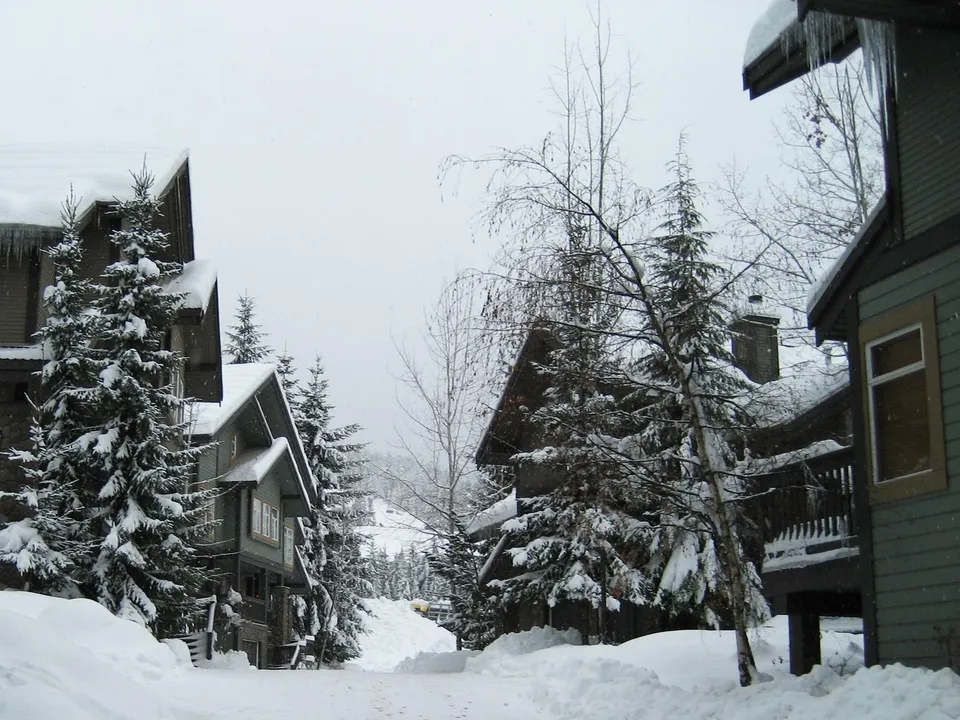 A village covered in snow with evergreen trees and houses under a cloudy sky.
