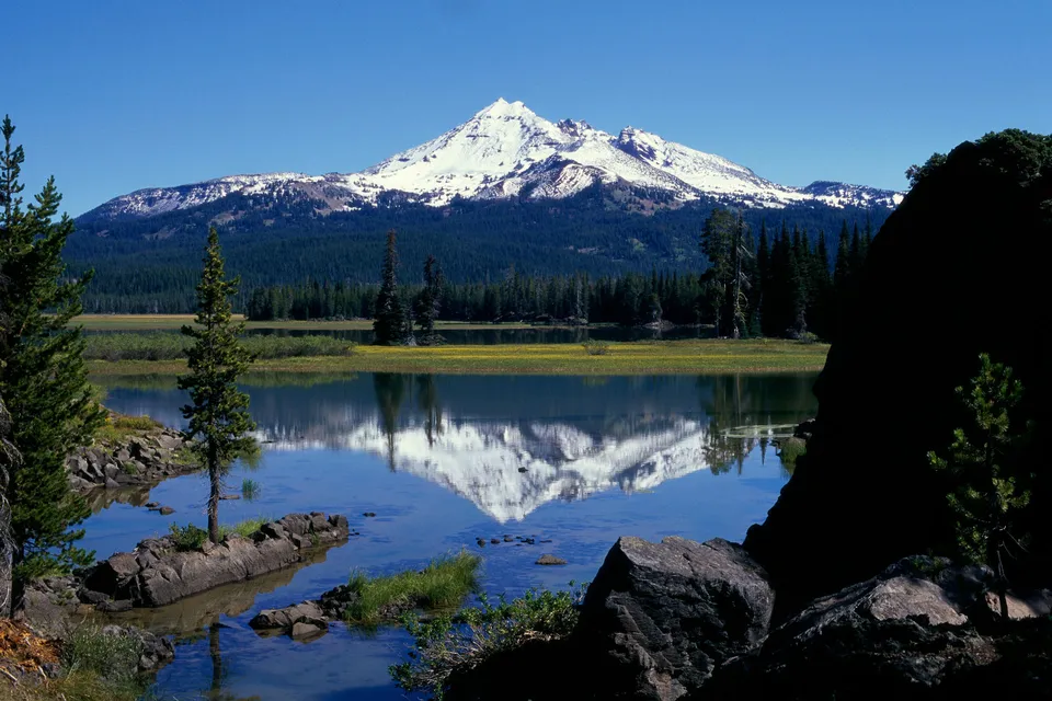 A snow-capped volcano is perfectly mirrored in the still blue water of a lake, with pine forests below.