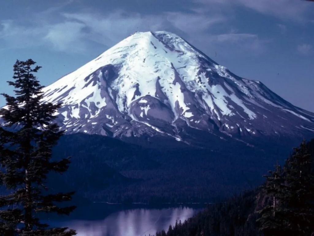 A snow-covered volcano rises above dark evergreen forests and a calm lake.