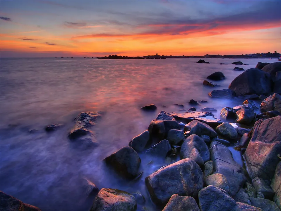 A serene sunset casts a soft glow over a rocky coastline and calm ocean.