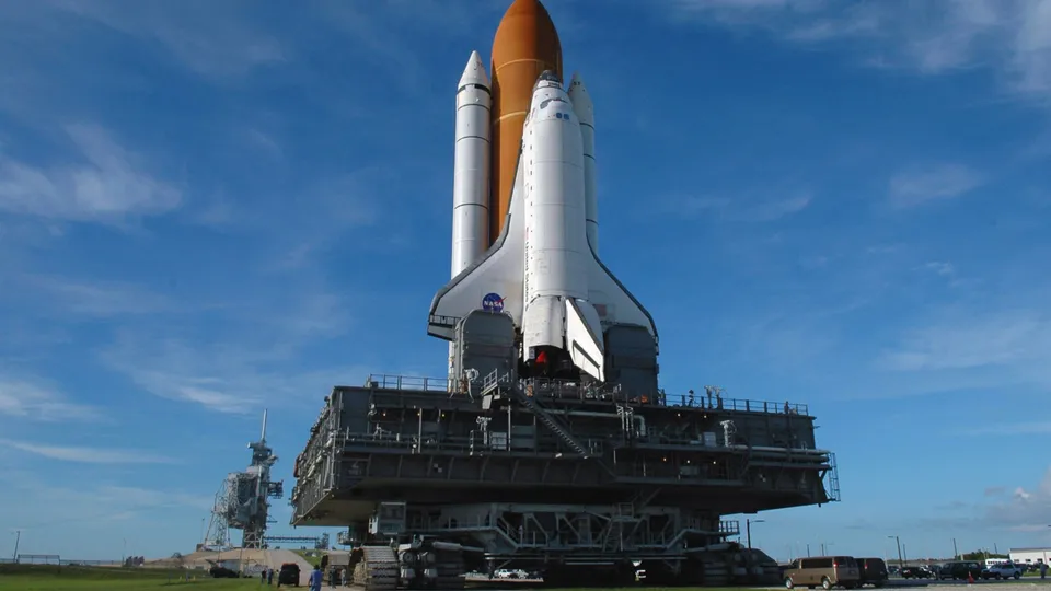 A space shuttle is positioned on a large crawler-transporter vehicle under a bright blue sky with scattered clouds.