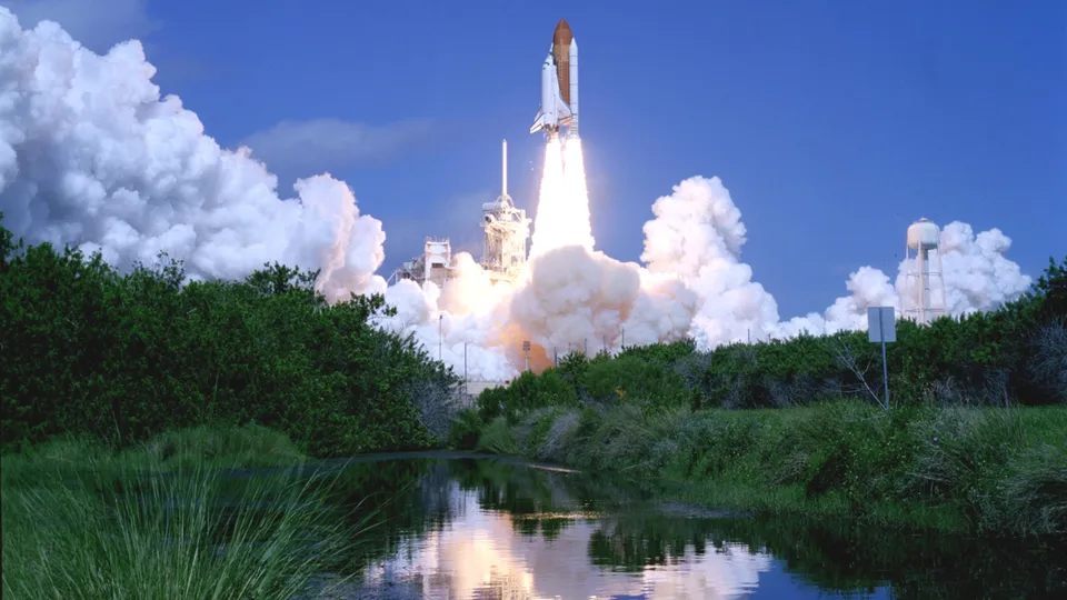 A space shuttle launches into a clear blue sky, trailing massive plumes of white smoke and fire.