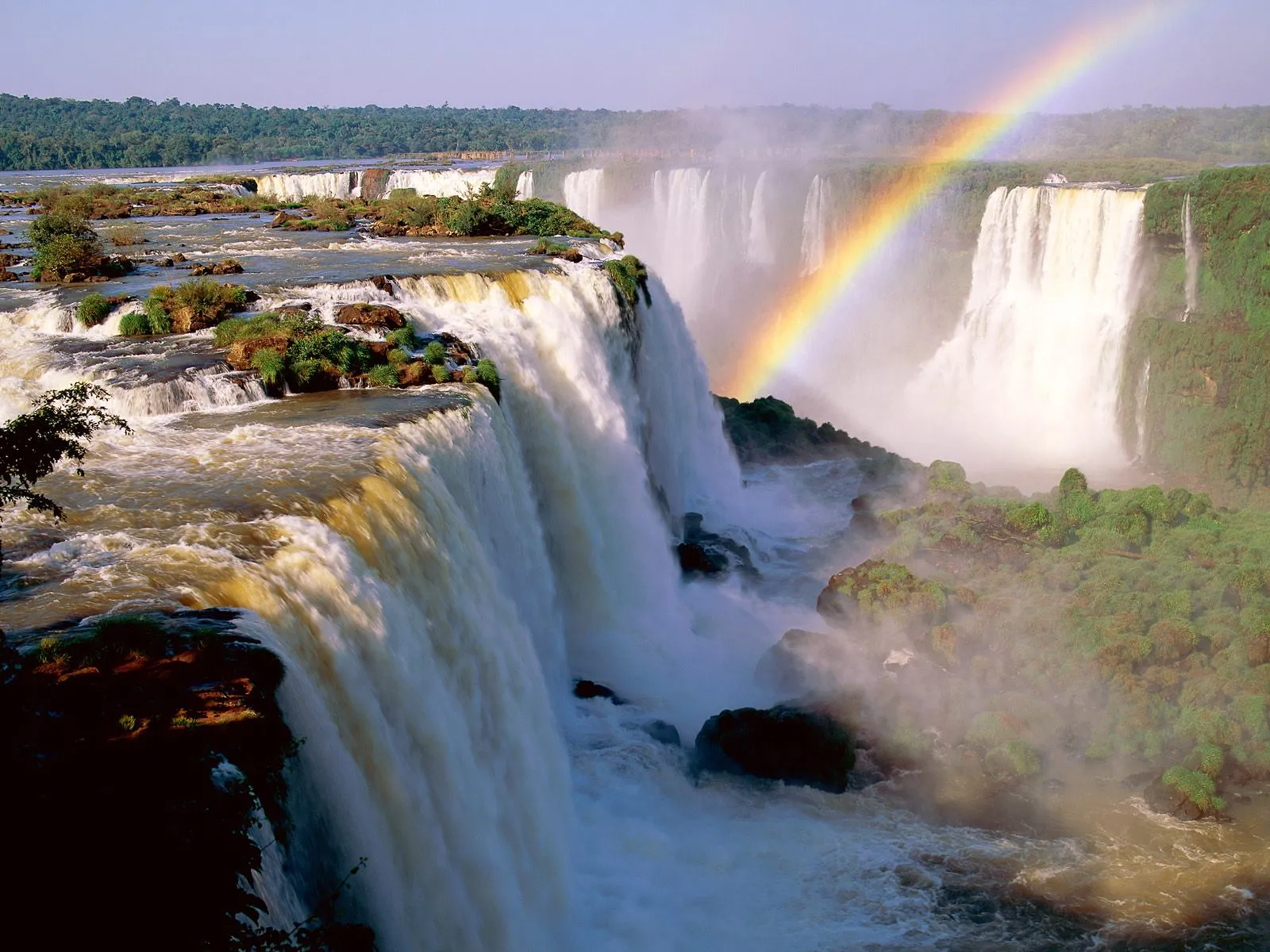 Numerous waterfalls cascade down lush green cliffs with a rainbow in the mist.