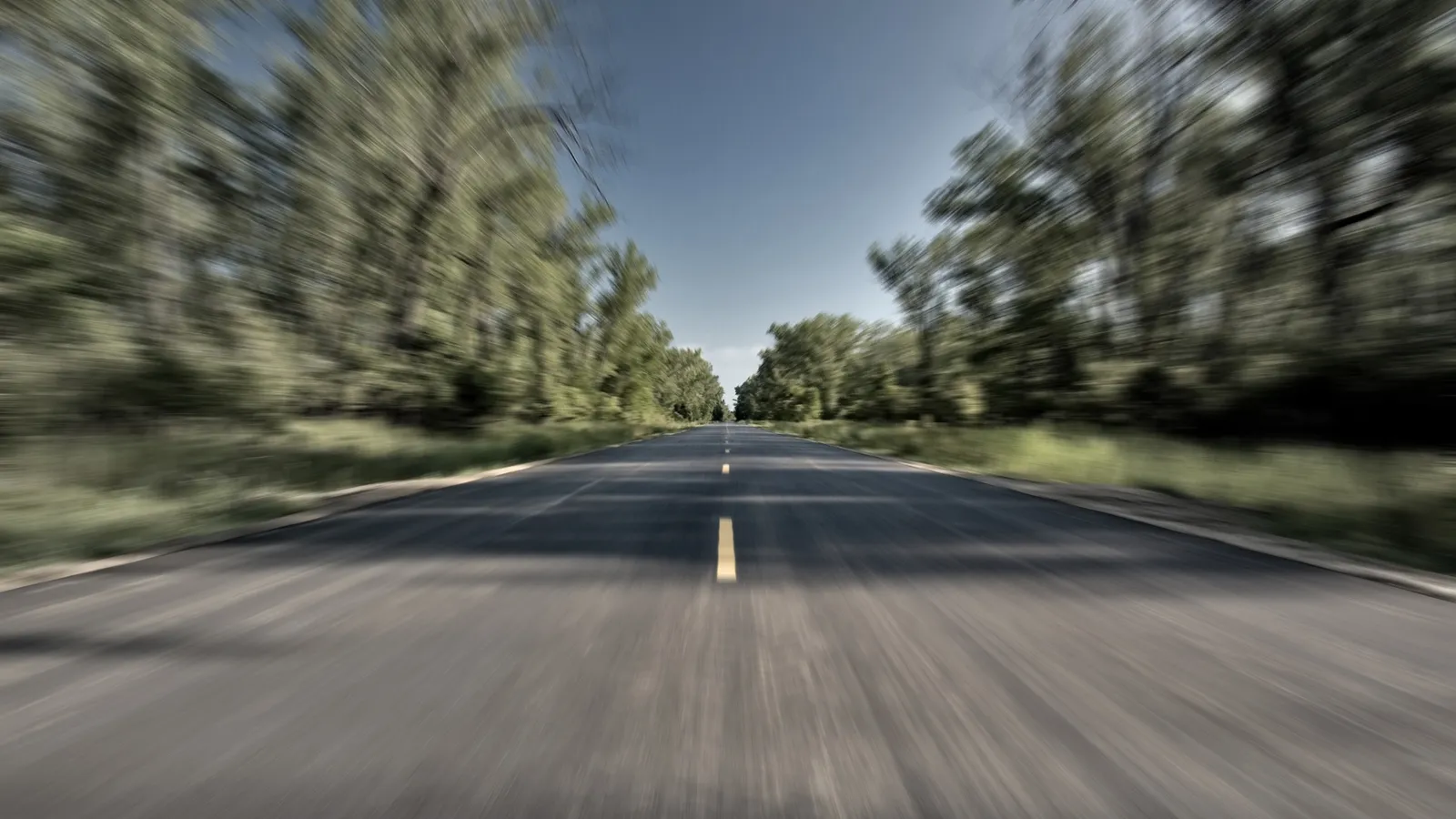 A blurred asphalt road stretches into the distance, flanked by trees on either side under a clear blue sky.